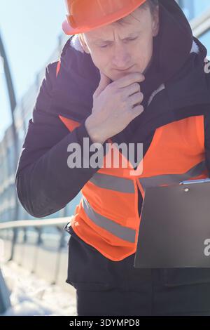 Male engineer in safety gear thoughtfully reviews documents outdoors in snowy urban environment Stock Photo