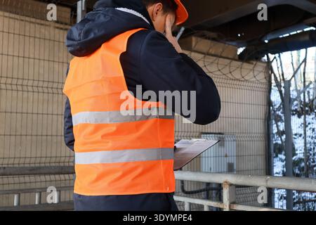 Side view of thoughtful worker in orange safety vest and hard hat reviewing documents on clipboard near fenced industrial zone Stock Photo