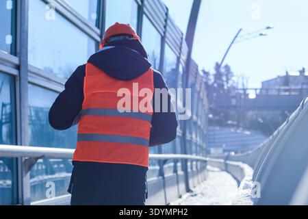 Young male in hard hat and vest stands on curved bridge path in sunny snowy cityscape Stock Photo