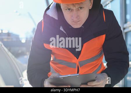 Close-up of male worker in safety attire intently studying clipboard documents at outdoor construction location in winter. Stock Photo