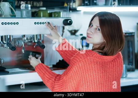 Wide angle of female worker in sweater smiling over shoulder as she presses controls on coffee equipment in cafe. Stock Photo