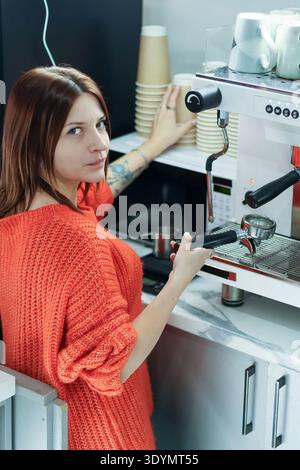 Side view of female cafe worker in orange sweater operating portafilter and reaching for items at espresso machine station. Stock Photo