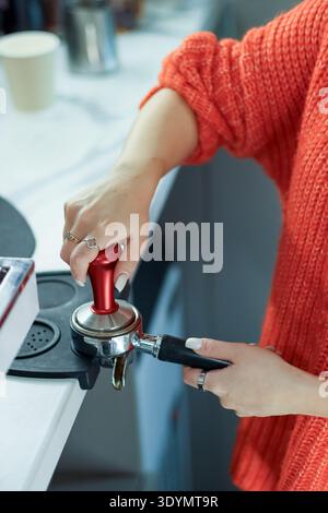 Vertical close-up of barista's hands pressing red tamper into coffee-filled portafilter on cafe counter. Stock Photo