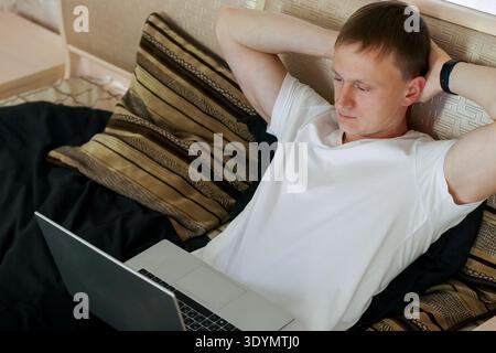 Casual young adult male resting arms behind head near open laptop on bed Stock Photo