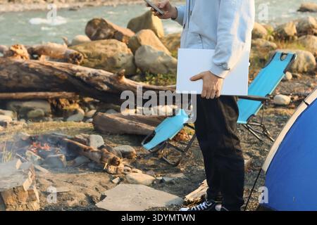 A young man stands casually holding a smartphone in one hand and a closed tablet in the other, next to a blue camp chair and tent by the rocky river. Stock Photo
