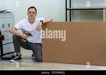 Man kneeling with drill leaning on large cardboard box in office environment. Focused man in casual attire squats holding drill, resting arm on tall c Stock Photo