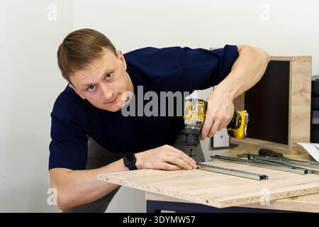 Man drilling into wood panel while assembling furniture. Focused man using yellow cordless drill on particleboard during furniture assembly at home Stock Photo