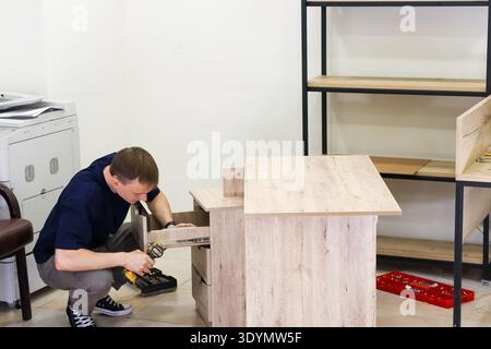 Carpenter installing drawer in light wood furniture. Skilled worker crouching and screwing drawer mechanism into wooden cabinet using power drill indo Stock Photo