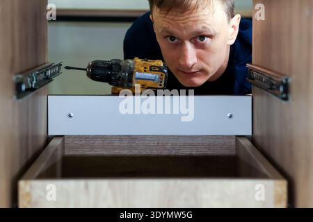 Worker drilling hole for drawer slide mounting. Close-up of craftsman using power drill to install metal drawer runner in wooden furniture piece. Stock Photo