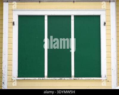 A close-up view of a yellow wooden house shows a large window with three panels, all of which are covered by solid dark green shutters Stock Photo