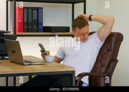 Pensive office worker pondering over smartphone screen with coffee cup during seated break in workspace. (92 characters) Stock Photo
