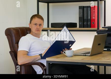 Serious young man intently studies paperwork inside blue folder in office setting, laptop displayed on desk with coffee mug adjacent. Stock Photo