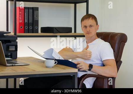 Young professional in office chair gives positive thumb gesture while reviewing blue file contents Stock Photo