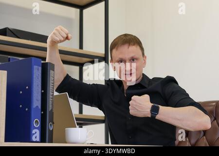 A young man in black shirt confidently flexes one arm while at laptop in office with binders and coffee. Stock Photo
