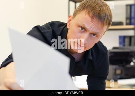 Man in office leans in to present two blank papers towards the camera, displaying a serious demeanor amid typical workspace elements. Stock Photo