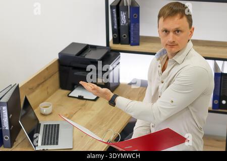 In office setting, male worker extends palm in gesture, clutching red folder near printer and laptop on wooden surface. Stock Photo