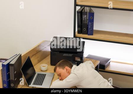 Stressed professional rests forehead on arms at desk, printer and laptop nearby, in bright office with document storage. Stock Photo