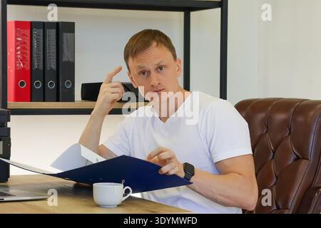 Reflective man tapping finger on temple while holding blue folder with papers at office setup Stock Photo