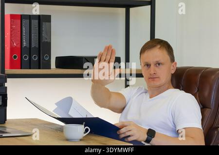 Young adult male in white shirt at office desk, hand raised palm out in halt signal, serious look, blue folder open with documents, cup on table, bind Stock Photo