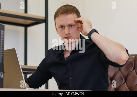 Professional appears perplexed, hand scratching head in confusion, seated before laptop in minimalistic office interior. Stock Photo