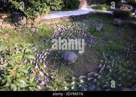 Intricate close-up of a spiral stone pattern in lush grass, framed by green shrubs, large rocks, and subtle garden paths for zen ambiance. Stock Photo