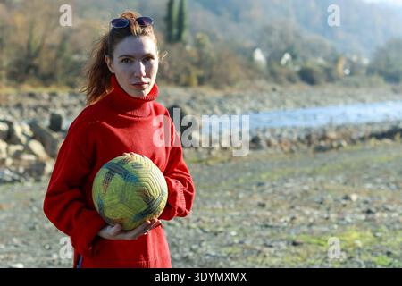 Young female in red turtleneck sweater holds yellow volleyball outdoors near river with misty mountains and rocky terrain in background. Stock Photo