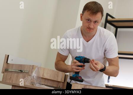 Young adult male in white shirt uses cordless drill to fasten screw into light wood drawer in bright indoor space. Stock Photo