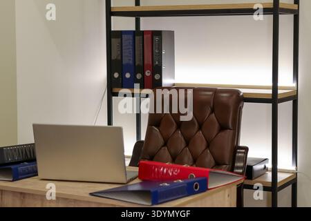 Modern office workspace featuring wooden desk, leather chair, open laptop, and colorful binders on metal shelf Stock Photo