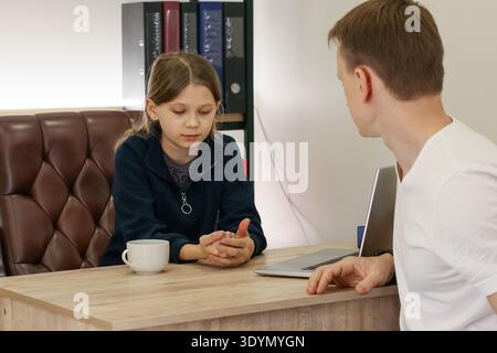 Child demonstrates with hands as adult indicates on closed laptop at desk with cup and office binders Stock Photo