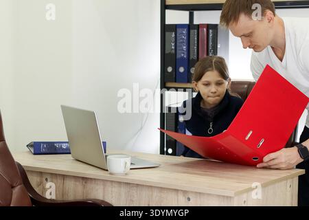 Adult male holds open red binder toward seated child at office table with laptop and cup nearby Stock Photo
