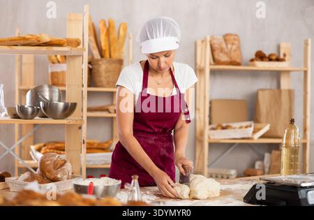 Focused young female baker in apron working at bakery, cutting dough into pieces Stock Photo
