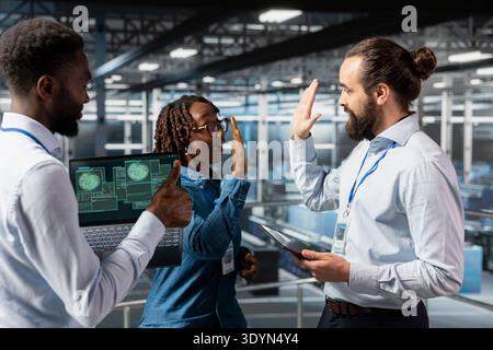 Diverse pleased staff sharing a high five to enjoy their accomplishment on server farm industrial platform, cheering to their professional collaboration. Happy successful engineering team. Stock Photo