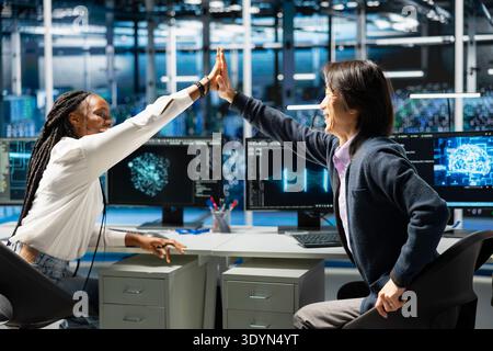 Delighted engineers celebrating in data center after successfully coding with generative AI. Jubilant coworkers doing high five after leveraging AI large language model to finish tasks Stock Photo