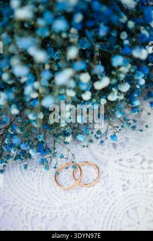 Gold wedding bands lie on a lace surface next to a delicate bouquet of small blue flowers captured during a wedding celebration in a bright setting Stock Photo