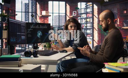 Man and woman watching business conference infographics on computer setup, looking over statistics on online portfolio. Analysts team review business presentation dashboard. Camera A. Stock Photo