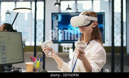 Female technician wearing VR goggles coding with AI language model assistance in startup office. Young woman using virtual reality integrating LLM assistant suggestions into code editor, camera A Stock Photo