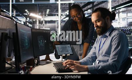Data center system administrators working together, collaborating on neural network software development. Server hub worker and african american colleague implementing AI tech in workplace, camera A Stock Photo