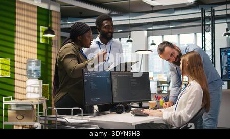 Friendly coworkers helping junior dev code AI algorithms to debug software issues and improve systems, teamwork concept. Welcoming team assisting newcomer in tech startup office, camera A Stock Photo