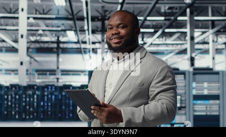 Engineer working in server farm using tablet to analyze data. IT professional examining infrastructure in data center using device, ensuring system integrity and security, camera B Stock Photo