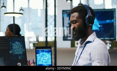 Computer scientist in office integrating AI machine learning systems to streamline workflows, earing headphones. African american man uses artificial intelligence on devices, listening music, camera A Stock Photo