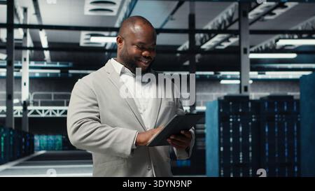 Smiling admin using tablet in in data center maintaining infrastructure for workloads. Joyful IT professional in server farm utilizing high tech server clusters for parallel processing, camera B Stock Photo