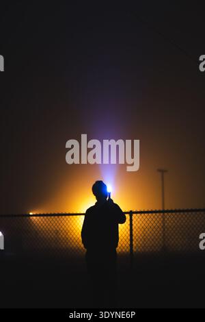 A person is standing in the dark with a flashlight in their hand, showing a silhouette in the bright yellow-orange light from a flashlight or torch. T Stock Photo