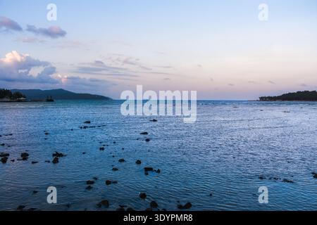 Calm coastal seascape at sunset featuring a rocky shoreline, gentle waves and distant hills beneath a pastel sky Stock Photo