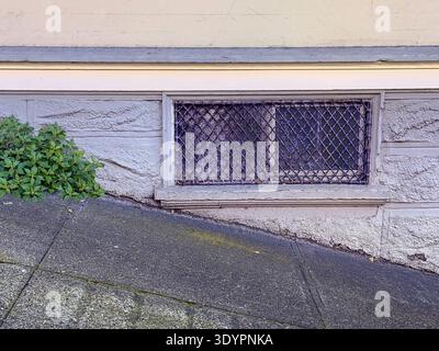A basement window covered by a decorative metal grate or vent cover with a diamond-patterned metal cover. The grate is installed on a light-colored st Stock Photo