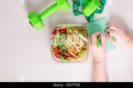 Healthy lunchbox with grilled chicken, lentils, fresh vegetable salad, and woman writing in notebook with dumbbells on desk Stock Photo