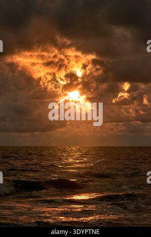 A stunning and dramatic sunset scene over the open ocean, featuring a vibrant, fiery sky with the sun breaking through dark, ominous clouds. The golde Stock Photo