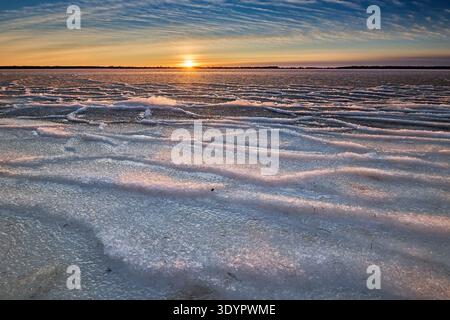 A wide-angle landscape photograph showcasing a frozen lake at sunset, with intricate rippled ice patterns and textured snow reflecting the warm orange Stock Photo