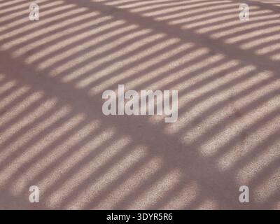 Sunlight casting strong, diagonal, parallel shadows across a warm, textured reddish brown ground surface, forming a repetitive striped pattern and pro Stock Photo