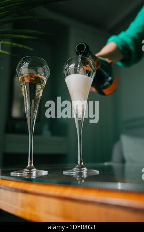 Hand pours sparkling wine into glass with sugar. Second glass filled with golden bubbly drink. Warm wooden bar surface. Cozy indoor lighting with gree Stock Photo