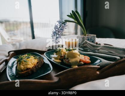 Two plates of breakfast food on wooden tray. One has avocado toast, the other eggs benedict. Small purple hyacinth flower beside drink. Soft light fro Stock Photo